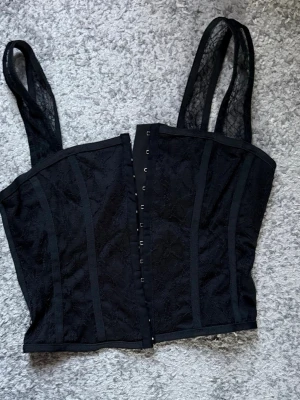 Svart spetskorsett från Missguided - Säljer en svart korsett från Missguided i storlek EUR 38. Toppen har breda axelband i spets, hakknäppning framtill och markerade sömmar för en snygg siluett. Materialet är spets med genomskinliga detaljer som ger en cool vibe.