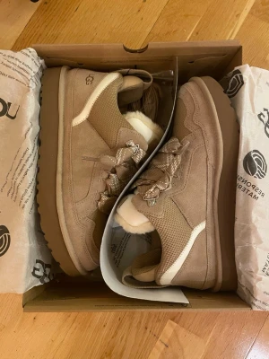 Beige sneakers från UGG med ullfoder - Säljer för att dom vart för stora. Säljer ett par snygga UGG Lowmel i färgen sand med breda, mönstrade snören och fluffigt vitt ullfoder. Ovandelen är i mocka och sulan är rejäl och greppvänlig. Perfekta för dig som vill ha både stil och komfort under höst och vinter.
