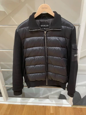 Svart pufferjacka från Bvaldi XS - Svart pufferjacka från Bvaldi i storlek XS. Jackan har quiltad framsida i syntetmaterial och stickade ärmar med en snygg patch på vänster arm. Dragkedja framtill och ribbad krage samt muddar. Perfekt för dig som gillar stilren och modern look.