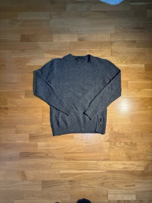 Grå stickad tröja Tiger of Sweden - Mysig grå stickad tröja från Tiger of Sweden, tillverkad av fin italiensk garn. Tröjan har rund hals och långa ärmar med ribbade muddar. Perfekt för kyliga dagar och enkel att matcha med jeans eller chinos.