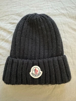 Moncler mössa - Säljer en svart ribbstickad mössa med uppvikt kant. Mössan är grovstickad och har en enkel, tidlös design som passar perfekt för kyliga dagar. Säljs då den inte används. 