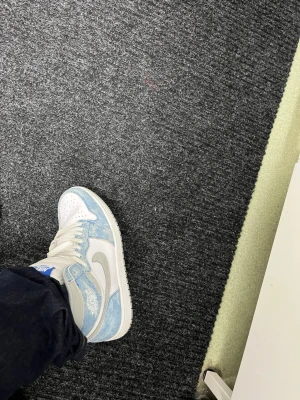 Nike Jordan 1 Hyper Royale  - Säljer ett par Nike Jordan 1 Hyper Royale i fräscht blått och vitt med grå detaljer. Skorna har snörning, rund tå och platt sula. Ovandelen är i mocka och skinn, med klassisk Air Jordan-logga på sidan. Perfekt för dig som vill ha en ikonisk och stilren sneaker.