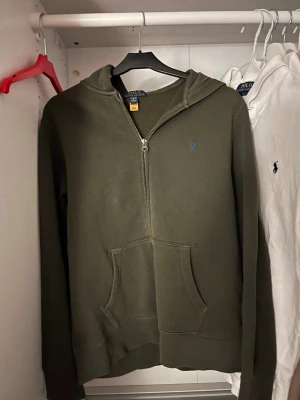 Militär/ mörk grön Ralph Lauren zip hoodie  - Militär/ mörk grön Ralph Lauren hoodie med dragkedja. Klassisk modell med broderad Polo-logga på bröstet och ficka framtill. Bra skick, inga hål eller större slitage. Skön och en stilren design som passar till vardags. Har ni frågor, prisförslag eller vill ha mer bilder skriv ett DM. Tröjan är i storlek L. OBS ❗️Detta är barn storlek, den är avsedd att passa juniorer inte vuxna. 