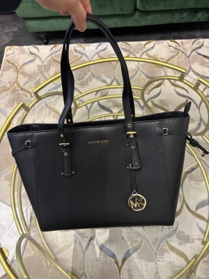 Svart handväska från Michael Kors - Stilren svart handväska från Michael Kors i skinn med guldfärgade metalldetaljer och MK-hänge. Väskan har två långa handtag och en klassisk, fyrkantig form. Perfekt för dig som gillar snygga och praktiska accessoarer med lyxig känsla. Kvitto finns. Väskan är köpt hos Michael kors i Japan Tokyo. Köptes som present men blev ändrade planer och nu säljs den. Hör av dig för fler foton eller andra frågor.