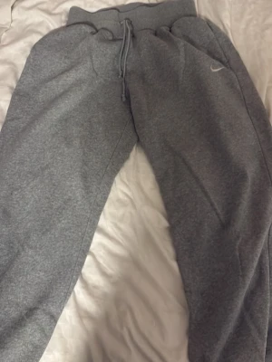 Grå mjukisbyxor från Nike - Säljer ett par grå mjukisbyxor från Nike med klassisk logga på benet. Byxorna har snörning i midjan och är tillverkade i mjukt bomullsmaterial. Perfekta för chill eller träning, med loose passform och elastisk midja för extra komfort. Ordinarie pris 750