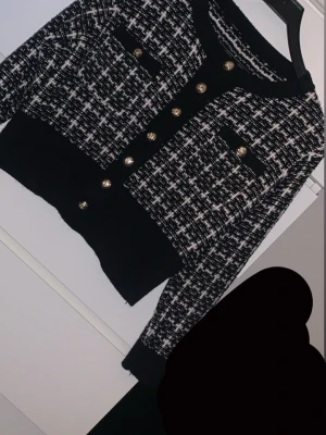 Svartvit rutig kofta med guldfärgade knappar - Säljer en snygg svartvit rutig kofta med guldfärgade knappar framtill och på fickorna. Koftan har lång ärm, rund hals och är croppad i modellen. Materialet är stickat och känns mjukt mot huden. Perfekt för dig som gillar klassisk stil med en modern twist.
