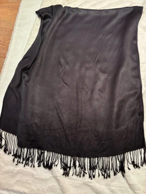 Svart sidensjal med fransar - Svart sjal i mjukt sidenliknande material med långa fransar längs nederkanten. Sjalen har en slät yta och små diskreta paljetter som ger lite extra glans. Perfekt för dig som gillar stilrena detaljer och vill ha något som sticker ut.