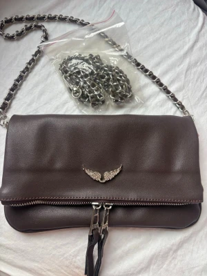 Brun axelväska från Zadig & Voltaire - Snygg brun axelväska i skinn från Zadig & Voltaire med silverfärgad kedja och dragkedja. Väskan har ett dekorativt emblem med vingar framtill och magnetlås. Insidan är fodrad med logotypmönster och har en innerficka. Perfekt för dig som gillar stilrena detaljer.
