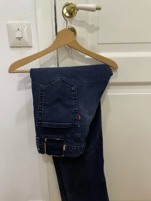 Levi's 501 mörkblå jeans med rak passform - Säljer ett par mörkblå jeans från Levi's med klassisk femficksdesign och rakt snitt. Jeansen har den ikoniska röda Levi's-taggen på bakfickan och är tillverkade i slitstarkt denim. Perfekta för dig som gillar tidlös stil och vill ha ett par jeans som funkar till allt.