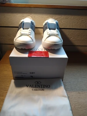 Valentino Garavani Open Sneakers, vit/blå - Säljer ett par Valentino Garavani Open Sneakers i vitt med marinblått band över mitten. Skorna har rund tå, platt sula och snörning. Tillverkade i skinn och levereras med originalkartong och dustbag. Perfekt för dig som gillar stilrena designer och lyxiga detaljer.
