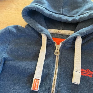 Blå Superdry hoodie med dragkedja - Blå hoodie från Superdry Orange Label med vit snörning och dragkedja framtill. Tröjan har huva, ribbade muddar och fickor på magen. Orange Superdry-logga på bröstet och detaljer på snörningen. Skriv vid minsta fundering