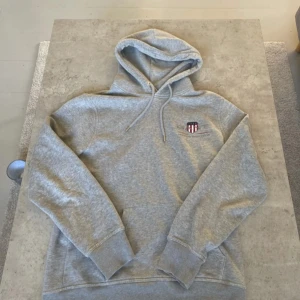 Grå hoodie från GANT  - Säljer en klassisk grå hoodie från GANT med broderad logga på bröstet. Tröjan har huva med snörning, magficka och mjukt material på insidan. Perfekt för chill dagar eller när du vill ha en enkel och snygg look.