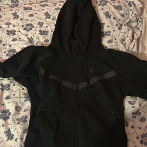 Svart Nike hoodie med dragkedja XS - Svart hoodie från Nike i storlek XS med hel dragkedja framtill och huva. Snygga svarta detaljer över bröstet och två fickor. Perfekt för chill eller träning, klassisk och stilren design.