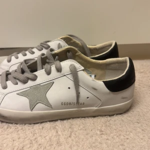 Golden Goose vita sneakers med stjärna - Golden Goose sneakers i vitt skinn med grå mockadetaljer och grå snörning. Sidan pryds av en ikonisk stjärna i mocka och baksidan har svart hälkappa med guldfärgad logga. Platt sula och rund tå ger en klassisk streetstil.