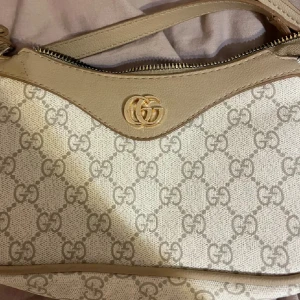 Beige axelväska GG-mönster - Snygg beige axelväska med klassiskt GG-monogram över hela väskan. Väskan har guldfärgad logga framtill, dragkedja upptill och detaljer i skinn. Perfekt för dig som vill ha en stilren och lyxig accessoar.