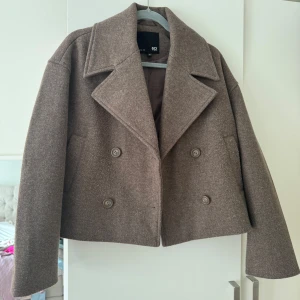 Brun skepparjacka från 157 - Säljer en brun skepparjacka från 157 med dubbelknäppning och bred krage. Jackan är croppad och har en loose passform med långa ärmar. Perfekt för dig som gillar klassisk stil med modern twist.