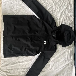 Svart vindjacka från Under Armour - Svart vindjacka från Under Armour med huva och hel dragkedja framtill. Fins i L och M. Jackan har en vit logga på bröstet och vita detaljer längs ärmarna. Tillverkad i lätt polyester, perfekt för blåsiga dagar. Snygg och sportig look med normal passform.
