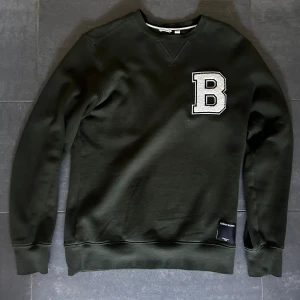 Svart sweatshirt från Björn Borg - Cool mörk grön sweatshirt från Björn Borg med stor vit B-applikation på bröstet. Tröjan har rund hals, långa ärmar och ribbade muddar. Perfekt för dig som gillar en stilren och sportig look. Nu pris 900kr