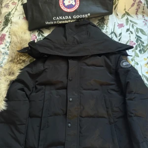  Canada Goose jacka - Säljer en svart jacka från Canada Goose med avtagbar huva och snygg pälskant. Jackan har klassisk patch på ärmen, knäppning med både dragkedja och knappar samt två fickor framtill. Perfekt för kalla vinterdagar och riktigt stilren look.