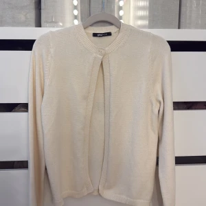 Beige kofta från Gina Tricot - En stilren beige kofta från Gina Tricot med rund halsringning och knapp framtill. Koftan är långärmad och har ribbade muddar vid ärmslut och nederkant. Perfekt att slänga över en topp för en enkel och clean look.