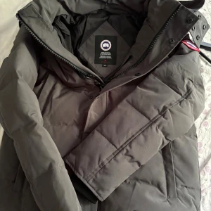 Grå Wyndham Parka från Canada Goose - Säljer en grå Wyndham Parka från Canada Goose med klassisk patch på ärmen. Jackan är dunfodrad, har huva och dragkedja framtill. Perfekt för kalla vinterdagar och har en stilren, rak passform. Märke och modell syns tydligt på innerlappen och etiketten.