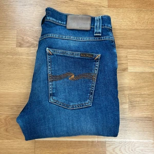 Nudie Jeans blå slim fit jeans - Säljer ett par klassiska blå jeans från Nudie Jeans med snyggt slitna detaljer och orange sömmar på bakfickorna. Modellen är slim fit med normal midja och raka ben. Jeansen är tillverkade i mjuk bomull med en skön stretch för extra komfort.
