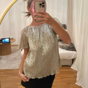 Zara topp - Från Zara. Passar en XS.Supersnygg glittrig blus i silver med korta ärmar. Blusen har ett plisserat tyg som ger en lyxig känsla och faller snyggt. Lös passform och vågig nederkant.