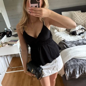 Svart omlott linnetopp med smock - Säljer en svart omlott linnetopp med smala axelband och smockad midja. Toppen har djup v-ringning och ett flowy fall nedtill. Perfekt att styla med kjol eller jeans för en trendig look.