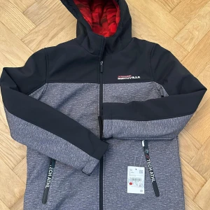 Grå och svart jacka från Northville - Sportig jacka från Northville i grått och svart med huva och röd fodrad insida. Jackan har dragkedja framtill, två fickor med dragkedja och tryckt logga på bröstet. Perfekt för dig som gillar en clean och funktionell stil.