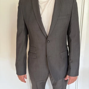 Grå kostym från Riley slim fit - Säljer en stilren grå kostym från Riley i slim fit-modell. Kavajen har två knappar, tre fickor med lock och en bröstficka. Fodret är mörkblått med ljusa detaljer. Använd typ 1 gång. Storlek 52.