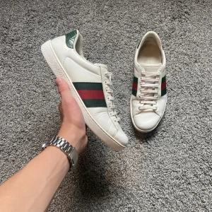 Gucci Ace vita sneakers med ränder - Gucci Ace sneakers i vitt skinn med klassiska gröna och röda ränder på sidorna. Skorna har snörning, rund tå och platt sula. Hälpartiet har en grön detalj med ormskinnsmönster. Perfekta för dig som vill ha en clean men lyxig look.
