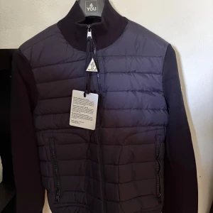 Moncler hybrid zip cardigan  - Snygg navy blue   quiltad jacka från Moncler med ribbad krage och ärmar. Jackan har dragkedja framtill, två fickor med dragkedja och Moncler-logga på bröstet. Framsidan är lätt vadderad och ger en modern look. Perfekt för dig som vill ha något stilrent och exklusivt.