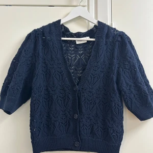 Mörkblå stickad kofta från Kappahl - Säljer en mörkblå stickad kofta från Kappahl i storlek S. Koftan har ett snyggt hålmönster, v-ringning och knappar framtill. Ärmarna är lite puffiga och slutar vid armbågen. Perfekt att slänga över en topp för en chill look.