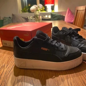 Svarta Puma sneakers med vit sula - Snygga svarta Puma sneakers i storlek 39 med vit, chunky sula och klassisk logga i guld på sidan. Skorna har snörning och är tillverkade i skinnliknande material med detaljer i textil. Perfekta för dig som gillar en stilren och sportig look.