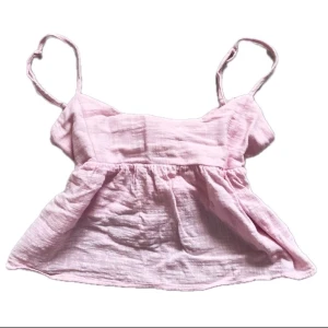 Ljusrosa linnetopp med rosett - Supersöt ljusrosa linnetopp med tunna axelband och en stor rosett i ryggen. Toppen är lätt utställd och har en luftig känsla, perfekt för varma dagar. Tillverkad i ett mjukt och svalt bomullstyg som ger en avslappnad vibe.