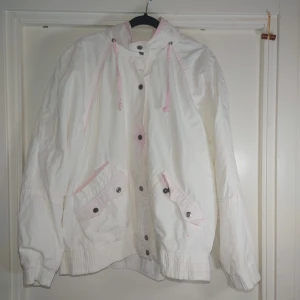 Vit vindjacka från New York Girl L - Vit vindjacka med ljusrosa detaljer från New York Girl. Jackan har dragsko i huvan, knappar och dragkedja framtill samt två stora fickor med lock och knapp. Lätt och luftig modell med resår i ärmslut och nederkant. Perfekt för vår eller höst.