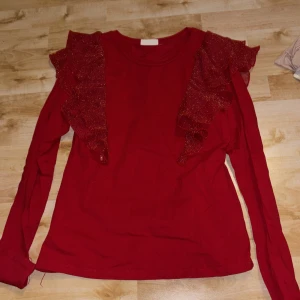 Röd långärmad topp med volang KappAhl - Snygg röd långärmad topp från KappAhl med glittriga volanger på axlarna. Perfekt för dig som vill sticka ut lite extra. Mjuk och skön bomullskänsla, rund halsringning och figurnära passform.