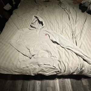 Grå hoodie från GANT med brodyr - Säljer en grå hoodie från GANT med broderad logga och sköld på bröstet. Tröjan har huva med snörning och klassisk känguruficka framtill. Mjuk och skön bomullsblandning, perfekt för chill dagar. Passar dig som gillar stilren och sportig look.