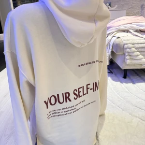Vit hoodie med text SELF-IMAGE - Vit hoodie med tryckt text 'SELF-IMAGE' i vinrött på både framsidan och baksidan. Tröjan har huva med snörning, magficka och ribbade muddar. Oversized passform och mjukt material som känns skönt mot huden. Perfekt för chill dagar. Passar bra som oversize modell.
