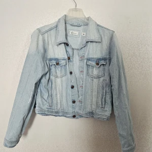 Ljusblå jeansjacka från Cdenim - Säljer en ljusblå jeansjacka från Cdenim i klassisk modell med krage, bröstfickor med knappar och långa ärmar. Jackan har snygga sömmar och bruna knappar framtill samt vid midjan. Perfekt för dig som gillar en chill och tidlös stil.