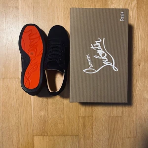 Christian Louboutin mörkblåa sneakers med kvitto - Säljer ett par svarta sneakers från Christian Louboutin med den ikoniska röda sulan. Skorna har snörning och är tillverkade i mocka med en stilren och klassisk design. Perfekta för dig som vill ha något exklusivt och snyggt i garderoben. Aldrig använda och kvittot kommer med. Tveka inte med frågor 