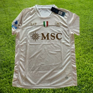 Napoli bortatröja 25/26 - Nyskick/helt oanvänd Napoli bortatröja från säsongen 202056/2026. Helt ärligt, detta är en av de vackraste tröjorna vi sett! Det vackra mönstret samt guldtrycken som glänser, wow! Tveka inte att höra av er för mer bilder och information!