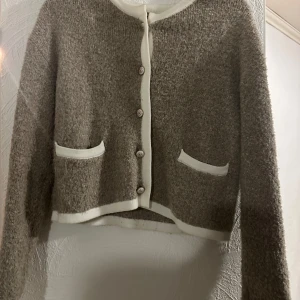 Beige stickad kofta med vita detaljer - Mysig beige stickad kofta med vita kanter och knappar framtill. Koftan har två fickor fram och är långärmad. Perfekt för lager-på-lager och chill dagar. Materialet är fluffigt och ger en soft känsla.