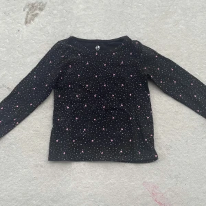Svart långärmad tröja med hjärtan H&M - Svart långärmad tröja från H&M i ekologisk bomull. Tröjan har ett gulligt mönster med små rosa hjärtan och grå prickar över hela tyget. Perfekt för dig som gillar söta detaljer och vill ha en bekväm basic med lite extra.