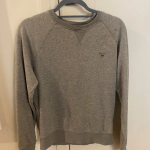 Grå sweatshirt från GANT storlek M - Grå sweatshirt från GANT med broderad logga och emblem på bröstet. Tröjan har rund hals, ribbade muddar och är tillverkad i mjuk bomull. Perfekt för chill dagar och enkel att matcha med jeans eller joggers. Helt ny använd bara 3-5gånger 