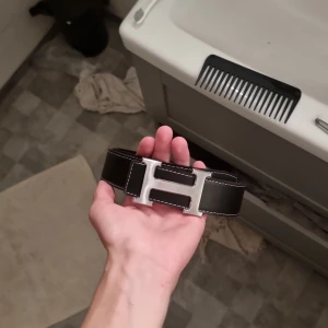 Svart skärp med H-spänne - Snyggt svart bälte med ett stort H-format metallspänne och vita kontrastsömmar. Bältet är gjort i skinn och har en stilren, klassisk look som passar till jeans eller kostymbyxor. Perfekt accessoar för att lyfta din outfit.