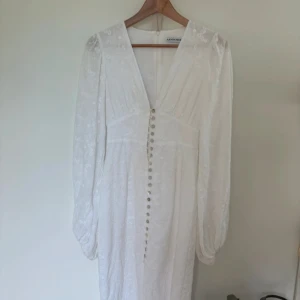 Vit broderad långklänning från Adoore - Superfin vit långklänning från Adoore med broderade blommor, v-ringning och långa ärmar. Klänningen har dekorativa knappar framtill och är tillverkad i ett lätt bomullstyg. Endast använd en gång , dessvärre saknas en knapp i övrigt superbra skick