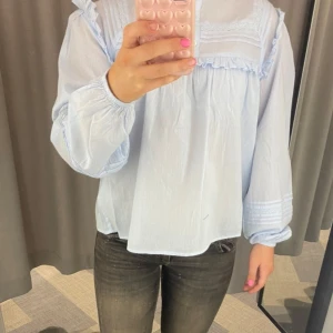 Ljusblå blus med volangdetaljer - Säljer en söt ljusblå blus med volangdetaljer och spets vid axlar och bröst. Blusen har långa ärmar med resår vid handlederna och en lös passform. Perfekt för dig som gillar romantisk och luftig stil.