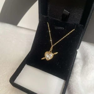 Guldigt halsband med hjärthänge - Supersött halsband i guldton med ett hjärtformat hänge som har en gnistrande sten i mitten och pil-detaljer på sidorna. Kedjan är tunn och elegant, perfekt för att ge din look lite extra shine. Hängsmycket har en romantisk vibe med hjärtmotiv. Rostfri och kjedjan är 40cm lång 