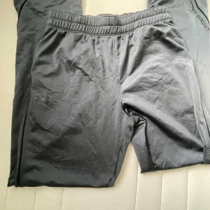 Svarta träningsbyxor från Under Armour - Svarta träningsbyxor från Under Armour i loose fit med resår i midjan och snörning. Byxorna är tillverkade i ett lätt och syntetiskt material, perfekta för träning. Diskret logga på benet och enkel, stilren design.